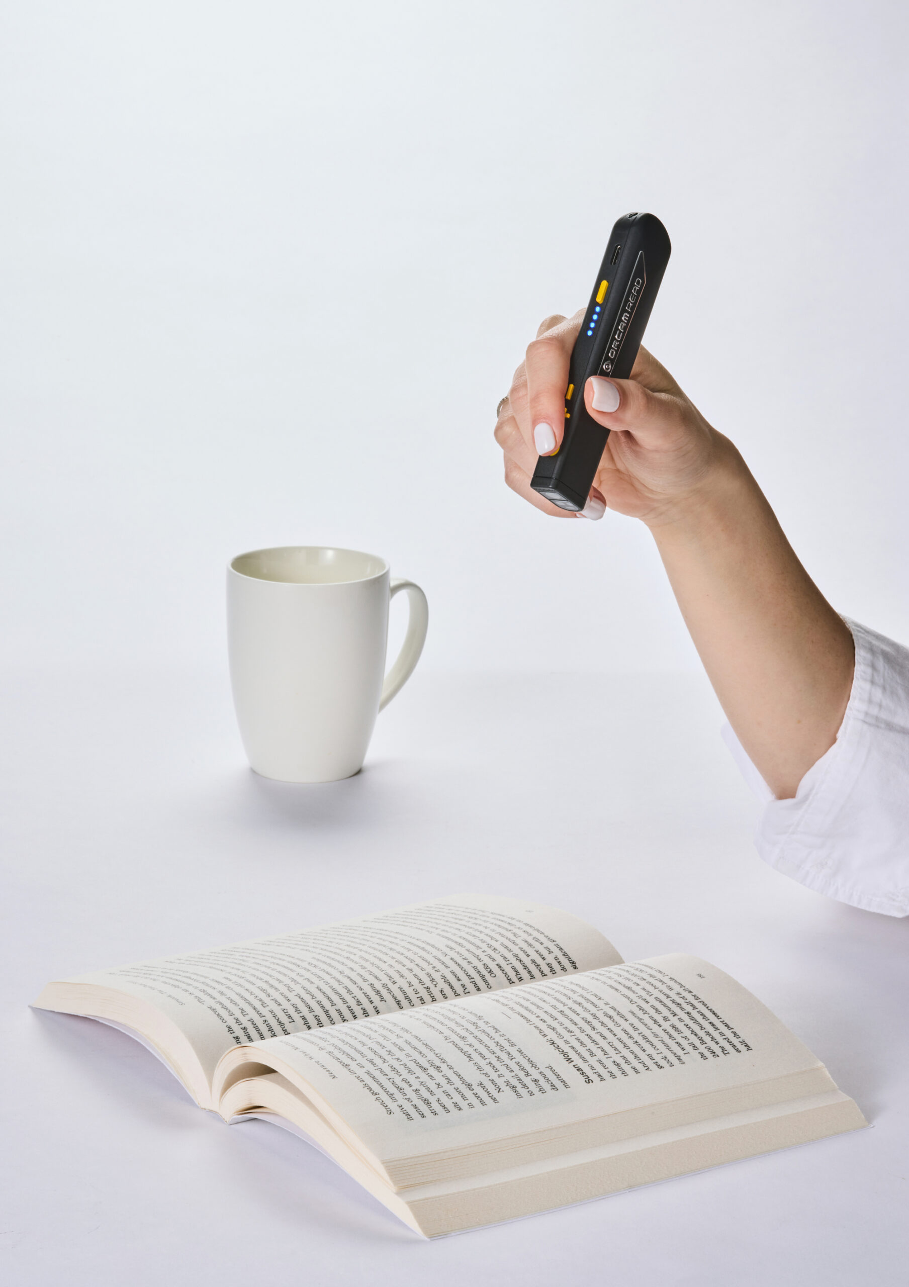 Billede af OrCam Read 3 som holdes over en åben bog, med et krus i baggrunden Billede af OrCam Read 3 som holdes over en åben bog, med et krus i baggrunden