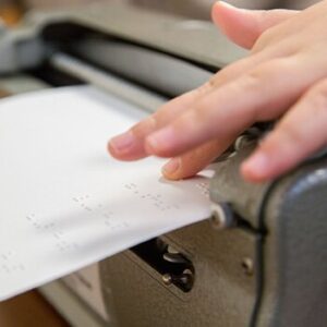 Billede af Perkins Brailler set bagfra, hvor en person berører papir på vej ud af enheden, med punktskrift på