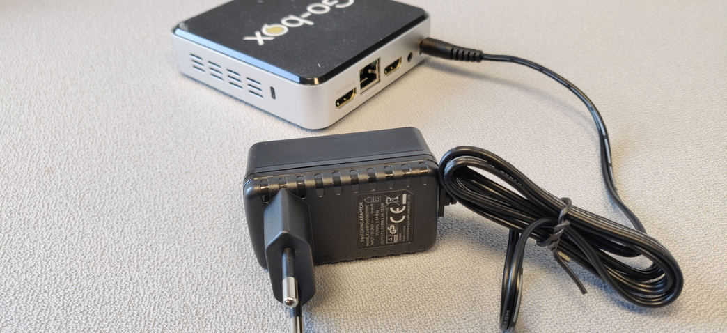 Billede af strømforsyning til Vo-box, Go-box samt Go-box Plus tilsluttet en Go-box i baggrunden